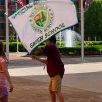 Student waves "Official Michigan Green School" flag in front of a plaza with a fountain